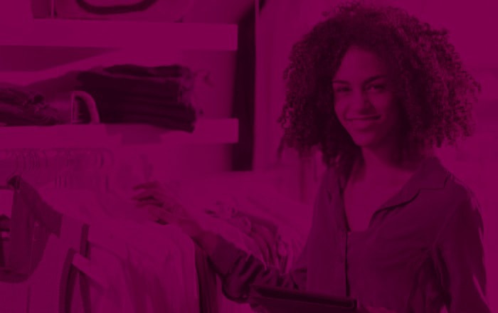 A person with curly hair smiles while looking at a rack of clothes in a store, holding a tablet.