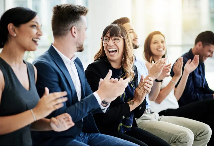 A group of six people sitting in a row, smiling and clapping during a gathering or event indoors.