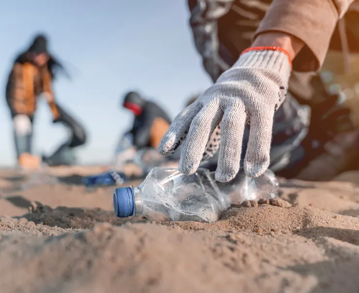 A gloved hand picks up a plastic bottle from the sandy beach while other people in the background participate in the cleanup.