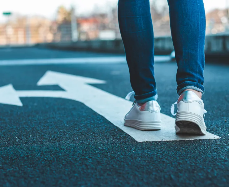 A person wearing white sneakers and blue jeans walks on an arrow-painted asphalt road, heading in the direction the arrow points.