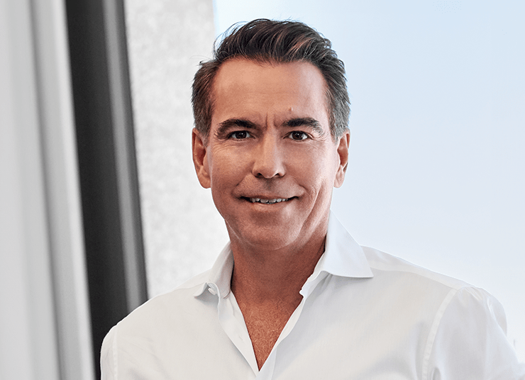 Headshot of Qlik Board of Directors member, Orlando Bravo.He has short dark hair, is wearing a white button-up shirt, and stands in front of a blurred background with a window and light blue sky visible.