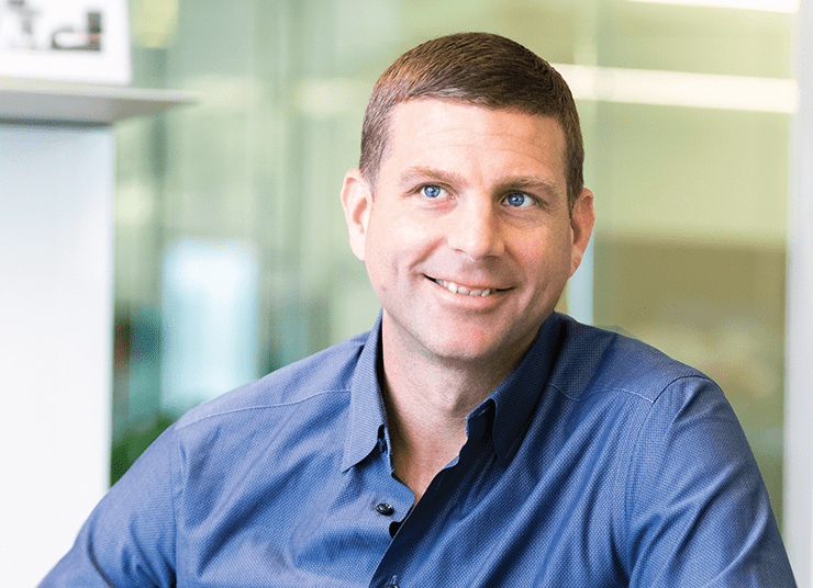 Headshot of Qlik Board of Directors member, Seth Boro. He is smiling, wearing a blue shirt, and sits in a brightly lit office setting.