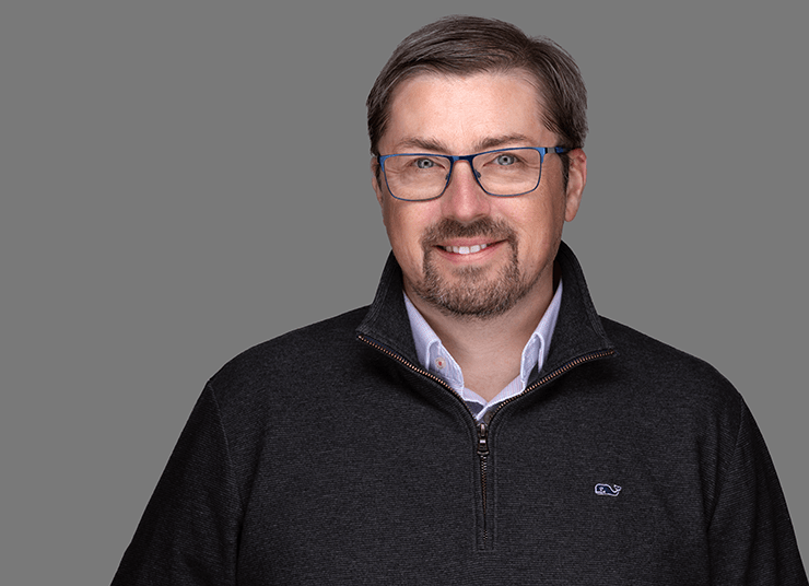 Brendan Grady, General Manager, Analytics Business Unit has short dark hair, glasses, and a beard smiles at the camera wearing a dark quarter-zip sweater and a collared shirt against a gray background.