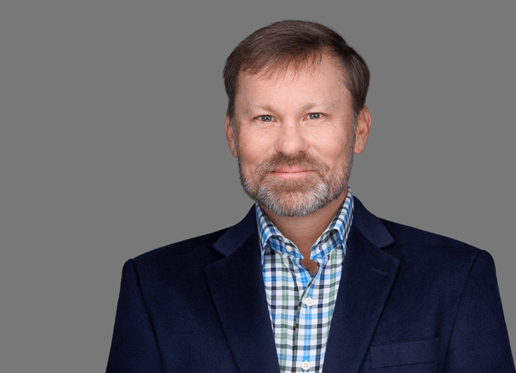 Casey George, Executive Vice President, Global Sales has short hair and a beard wearing a plaid shirt and dark blue blazer standing against a grey background.