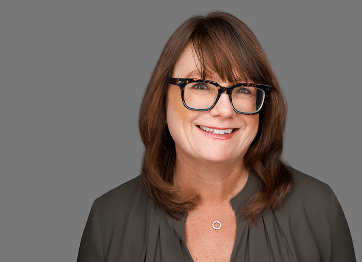 Deborah Lofton, Chief Legal Officer and Corporate Secretary with shoulder-length brown hair and glasses smiles at the camera against a plain gray background. She is wearing a dark blouse and a small pendant necklace.