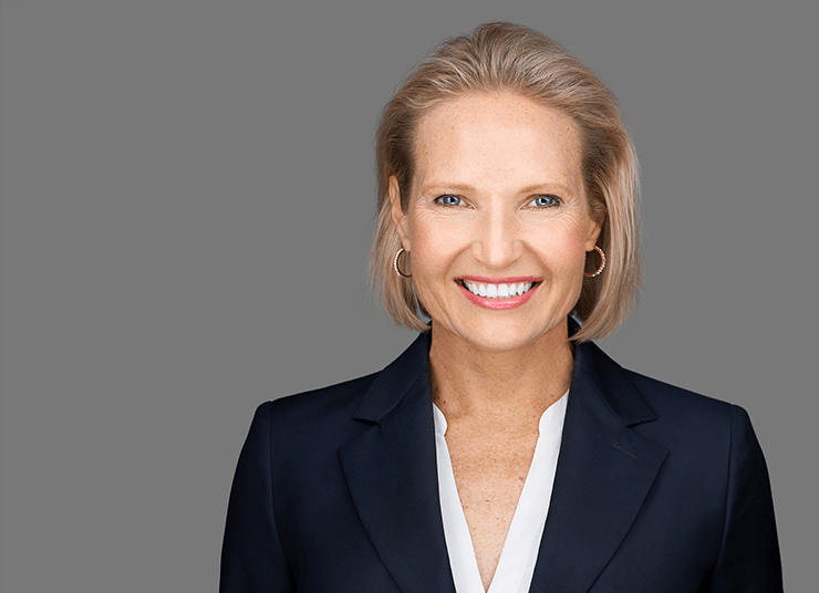 Julie Kae, VP, Sustainability and DE&I, Executive Director of Qlik.org with short, blonde hair and blue eyes is smiling while wearing a black blazer and white blouse against a gray background.