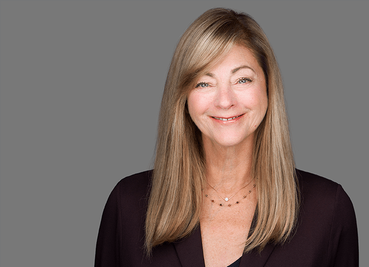 Ruthann Wry, Chief People Officer at Qlik has long blonde hair wearing a dark blazer and a necklace, smiling at the camera against a plain grey background.