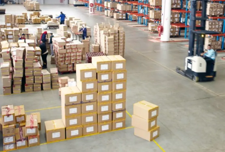 Warehouse filled with stacked cardboard boxes. Workers are organizing boxes and one is operating a forklift. Rows of shelving units with more boxes are visible in the background.
