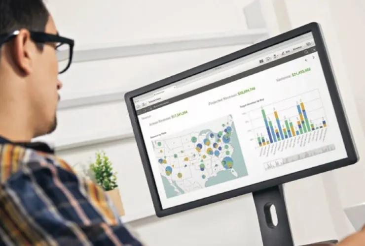 A person wearing glasses looks at a computer monitor displaying a U.S. map with data points and a bar graph with revenue figures.