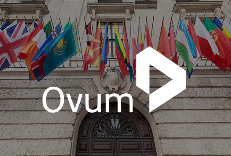 A building with multiple international flags displayed above a grand entrance. The word "Ovum" and a geometric logo are superimposed on the image.