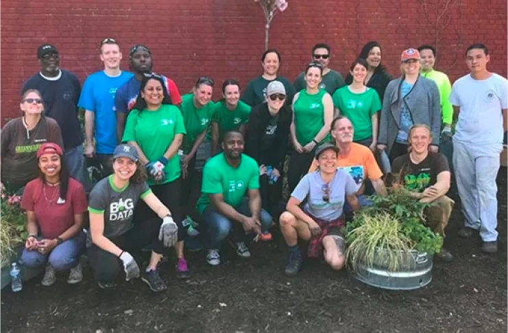 A group of 21 people are posing in front of a red brick wall. Some are standing, and some are kneeling, with plants in the foreground. They are dressed casually in T-shirts and working clothes.