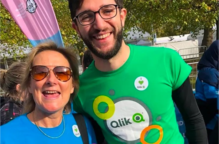 Two people standing outside, smiling at the camera. They are wearing Qlik branded shirts and have stickers with the same logo. Trees and event structures are visible in the background.