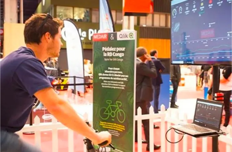 A man rides a stationary bicycle in front of a screen at an event, promoting a program labeled "Pédalez pour la RD Congo." The display tracks the cycling progress digitally.