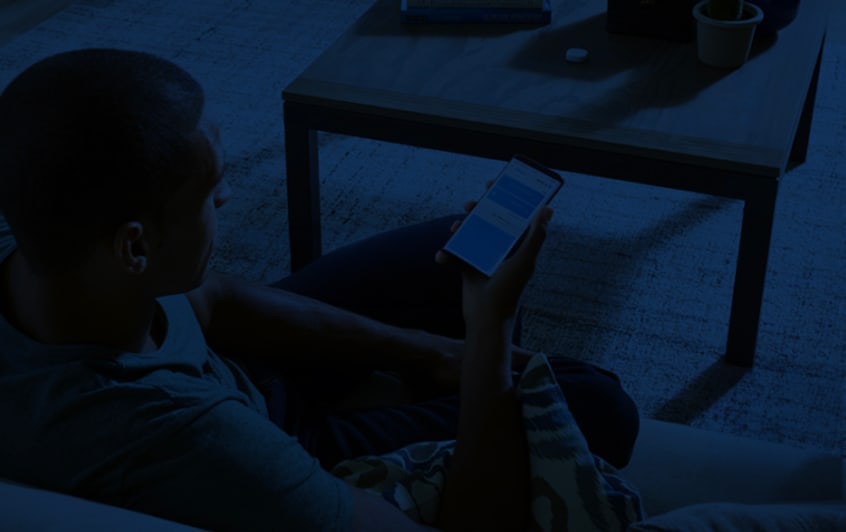 A person sitting on a couch looking at a smartphone screen in a dimly lit room with a wooden coffee table nearby.