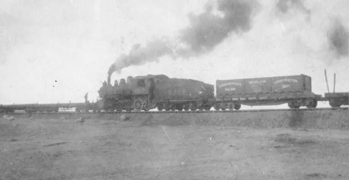 This black and white photograph shows a steam engine pushing flat cars with a man on it, behind the engine is the coal car and another car with Canadian Pacific Railway, Water Car, Construction Department and 10227 visible on it. Written on the back in pencil is “Ray Madden / First train to come to Shaunavon”