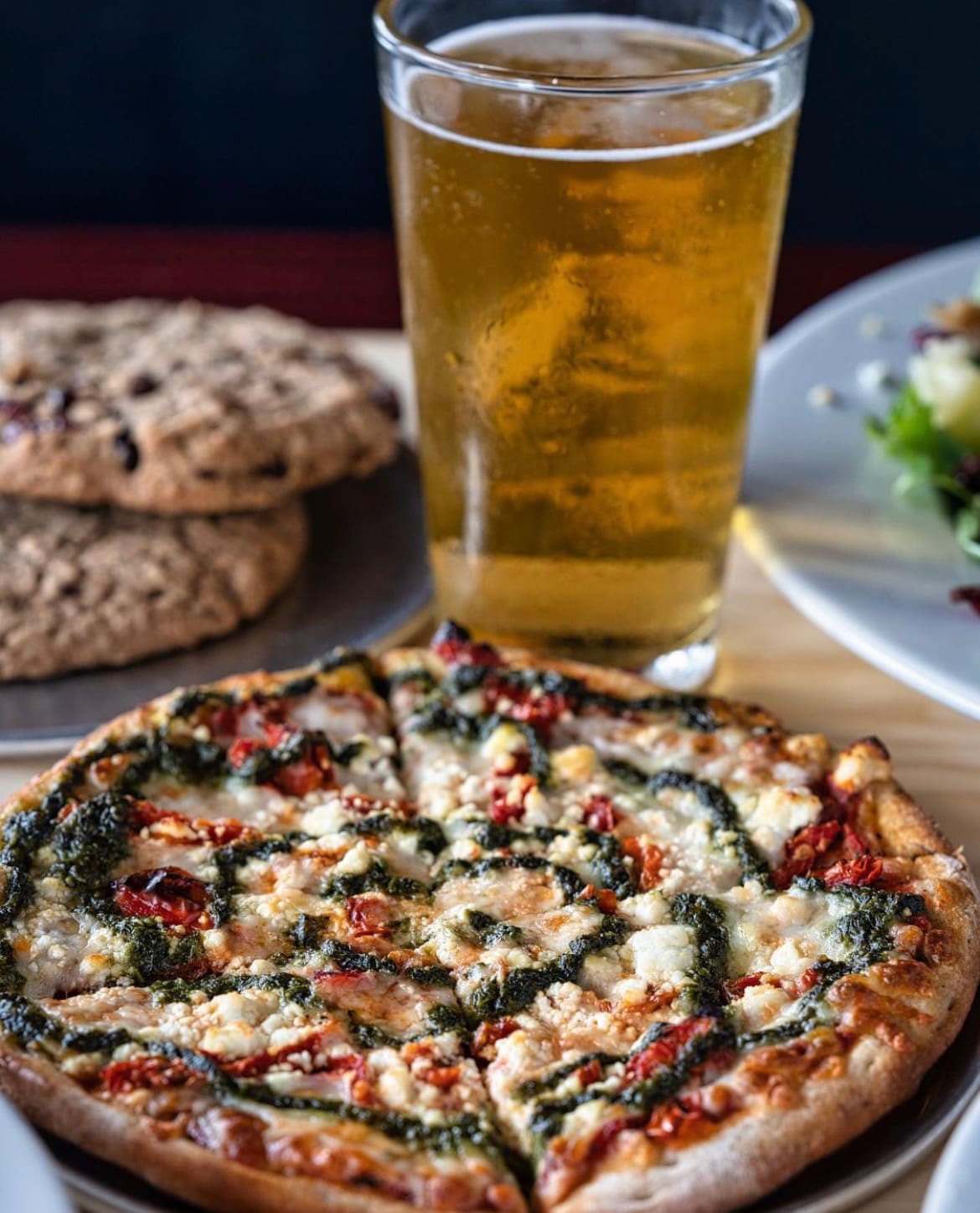 A pesto pizza, a cold pint of beer and cookies on a table at The Pizza Kitchen