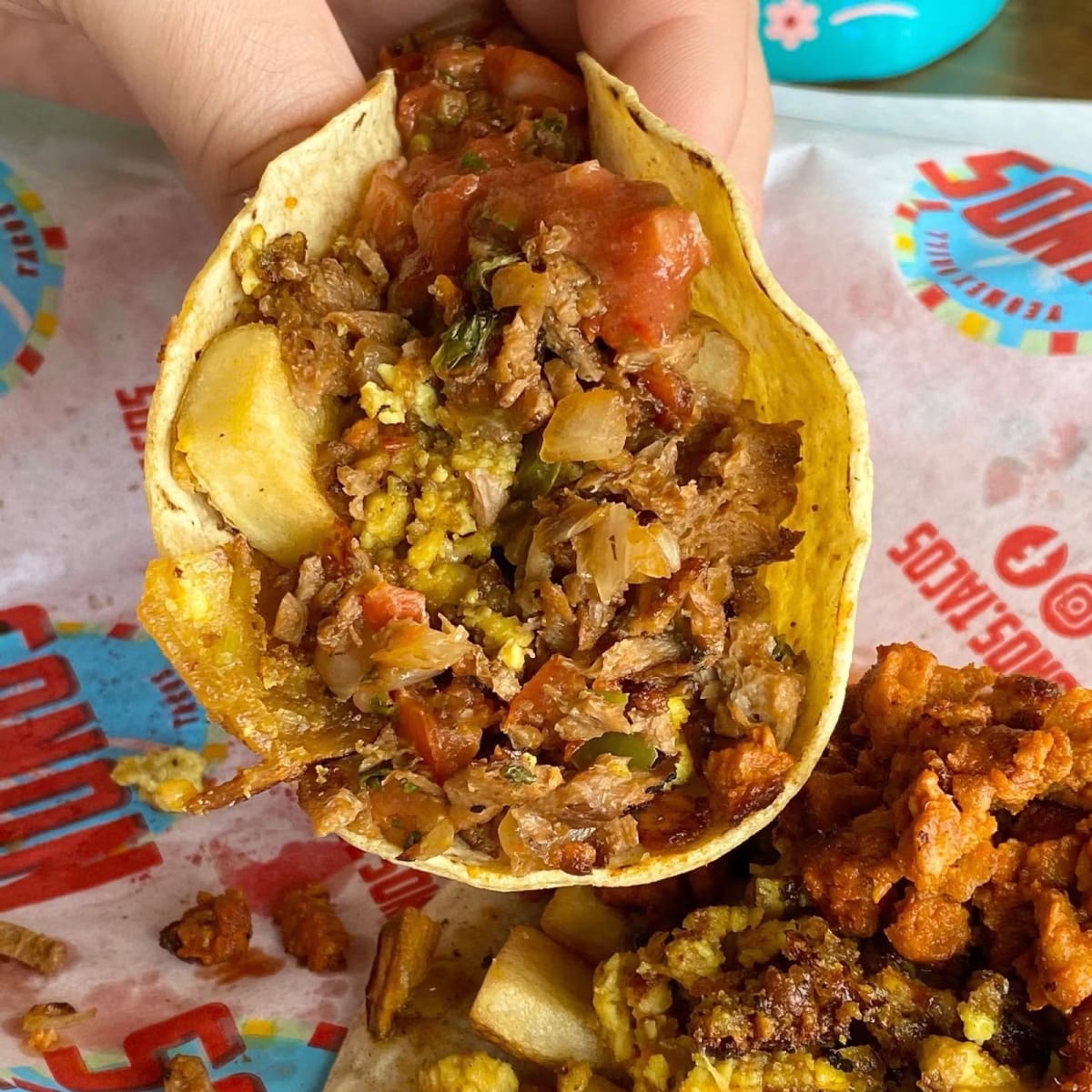 A loaded Nuno's taco held over branded takeout paper