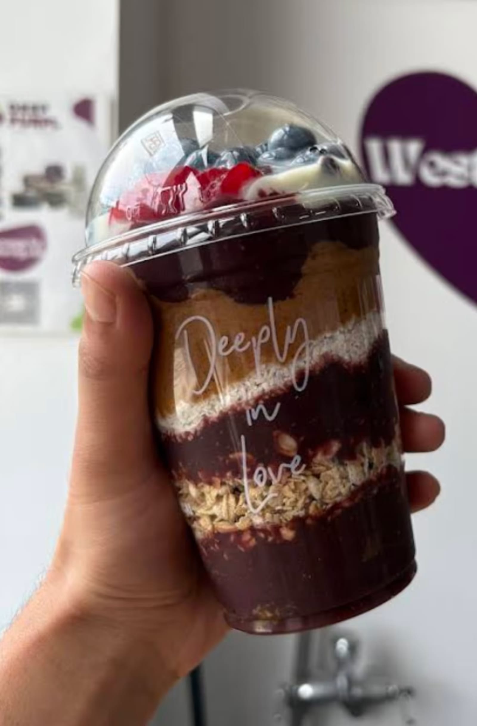 A hand holding a layered Deep Purpl açaí cup printed with Deeply in love