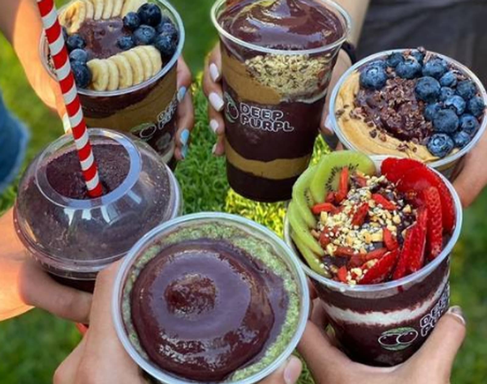 Three hands holding Deep Purpl açaí bowls and smoothies outdoors