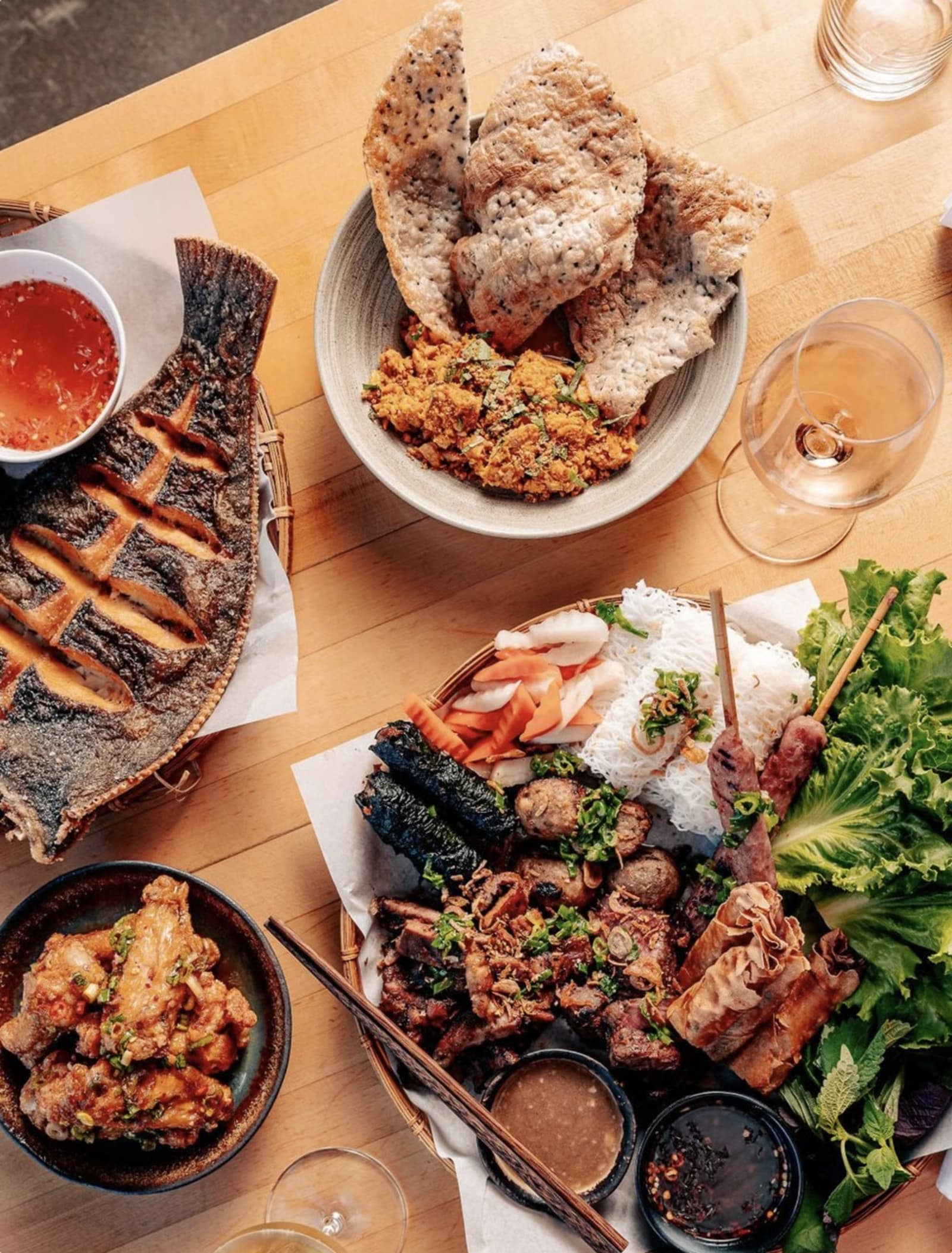 A family-style HaiSous spread — whole grilled fish, skewers, chicken wings, rice crackers, and fresh herbs