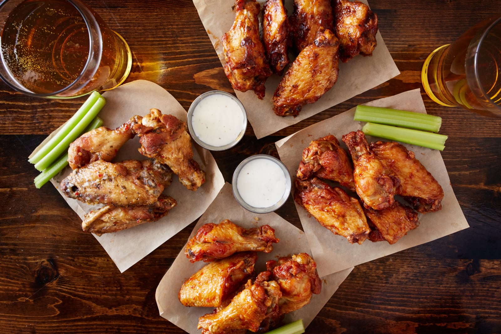 A board of chicken wings with celery, carrots and blue cheese dressing beside two glasses of beer