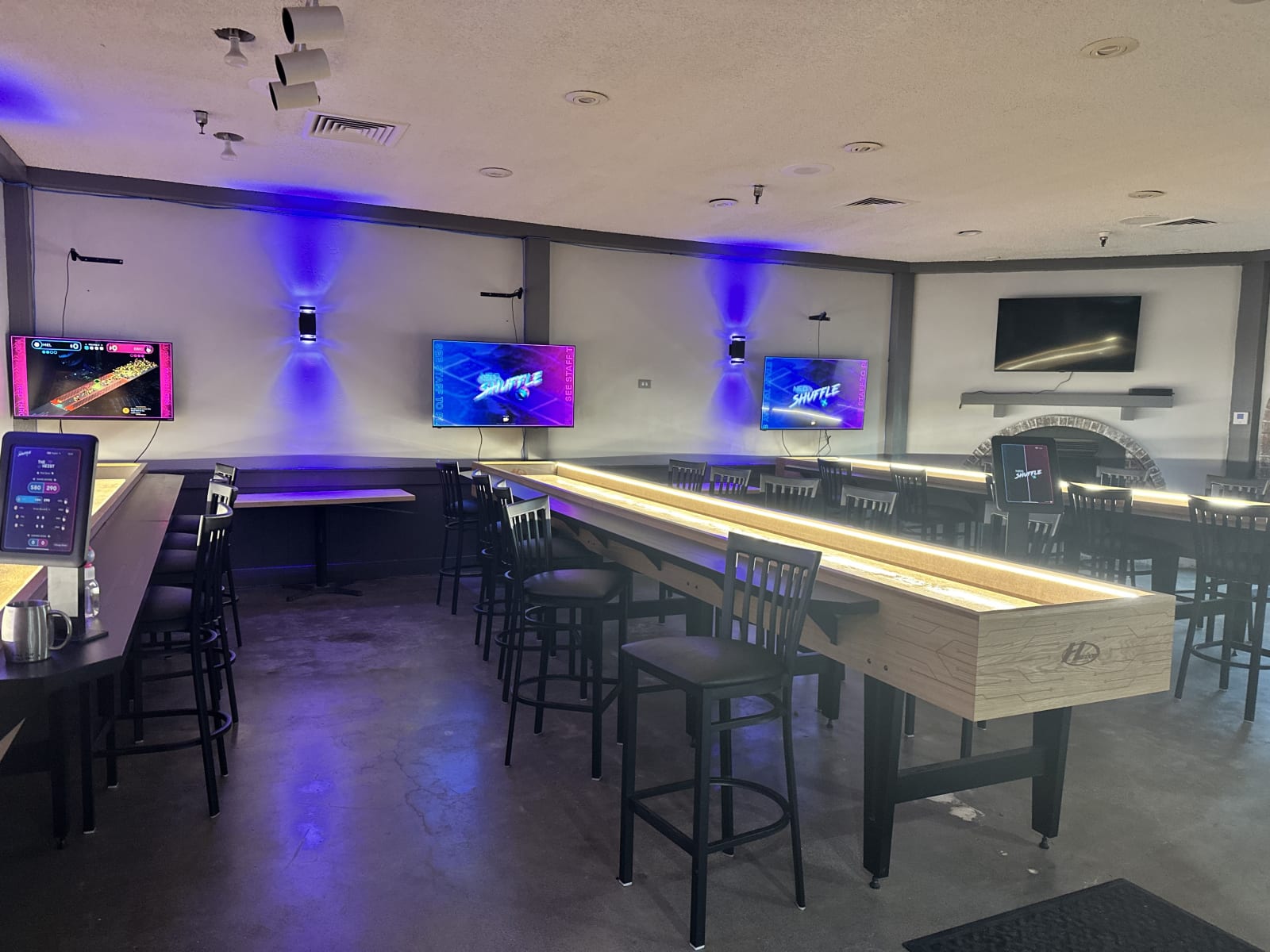 Two shuffleboard lanes and bar stools under purple wall-wash lighting
