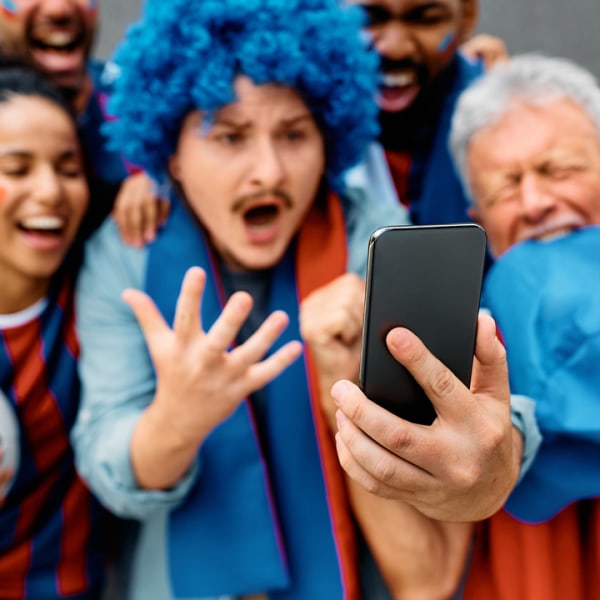Excited football fans in colourful wigs and scarves taking a selfie with a phone.