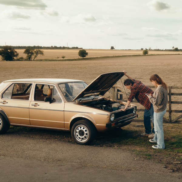 A couple inspects a car's engine on a rural road, with fields in the background.