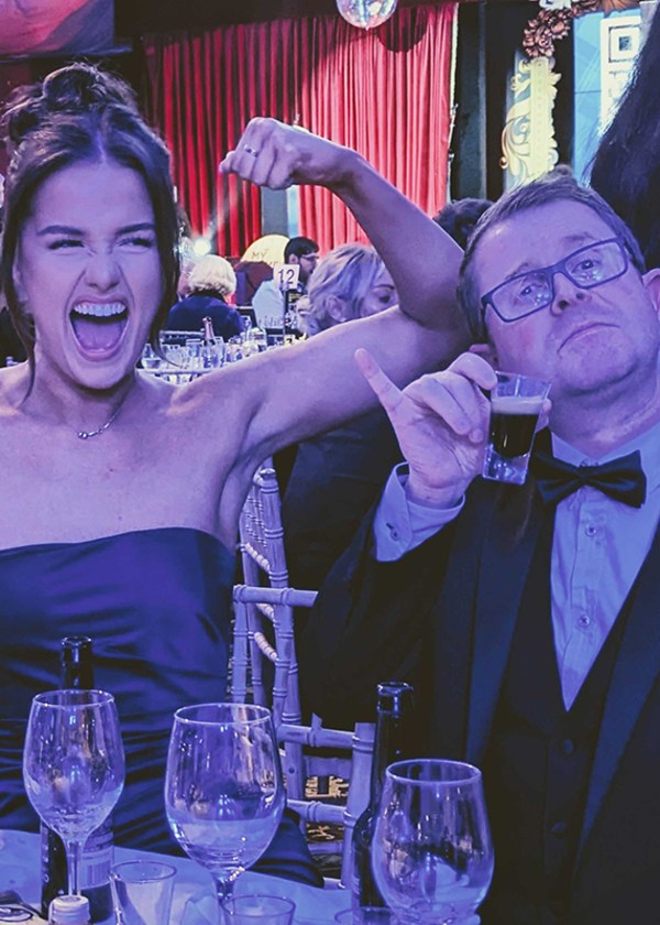 Woman flexing arm, man pointing at her, both smiling at a festive event.