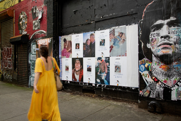 A woman in a yellow dress walks past posters and graffiti on a black wall.