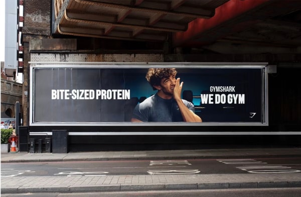 Billboard under a bridge with a man eating, text "BITE-SIZED PROTEIN" and "GYMSHARK WE DO GYM".