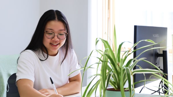 A smiling woman with glasses writing at a desk, beside a potted plant and a computer monitor.