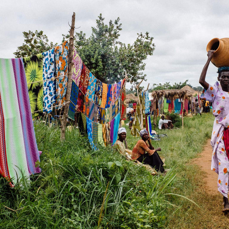 A community in Uganda where brightly coloured fabric is sold at the side of the road and a woman walks past carrying an earthenware pot.