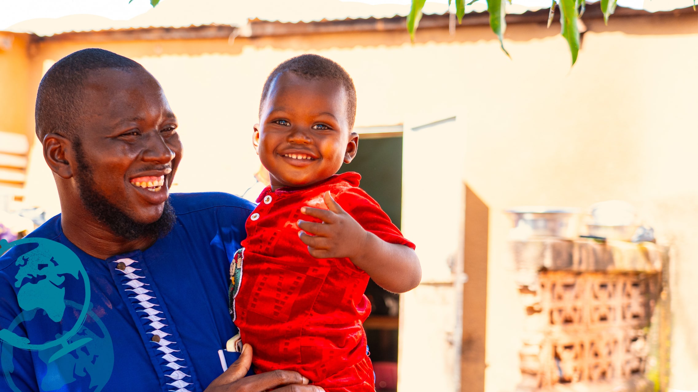 A man wearing a bright blue shirt holds a young child in his arms. They both smile to the side of the camera.