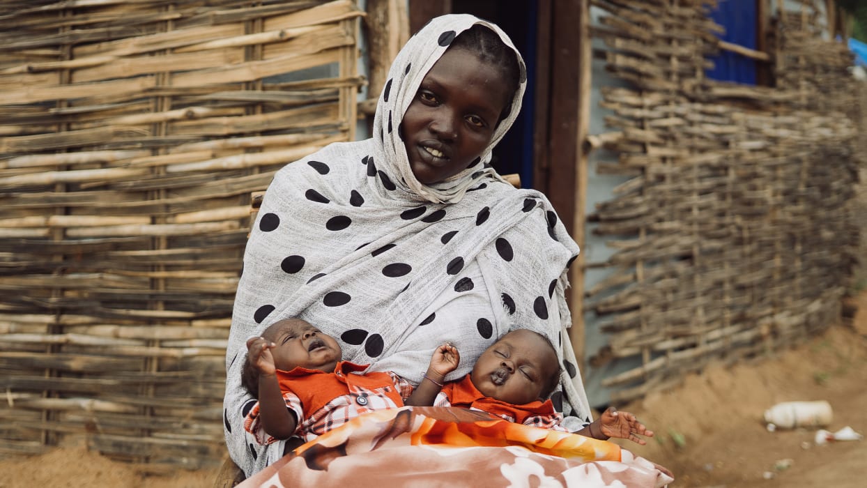 A woman in a white, black polka-dotted headscarf holds two babies. One baby is crying with open eyes, the other is sleeping. A woven wall forms the background.