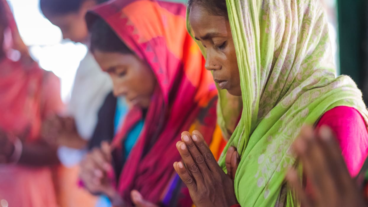 Women in colorful head coverings praying with clasped hands, eyes closed.
