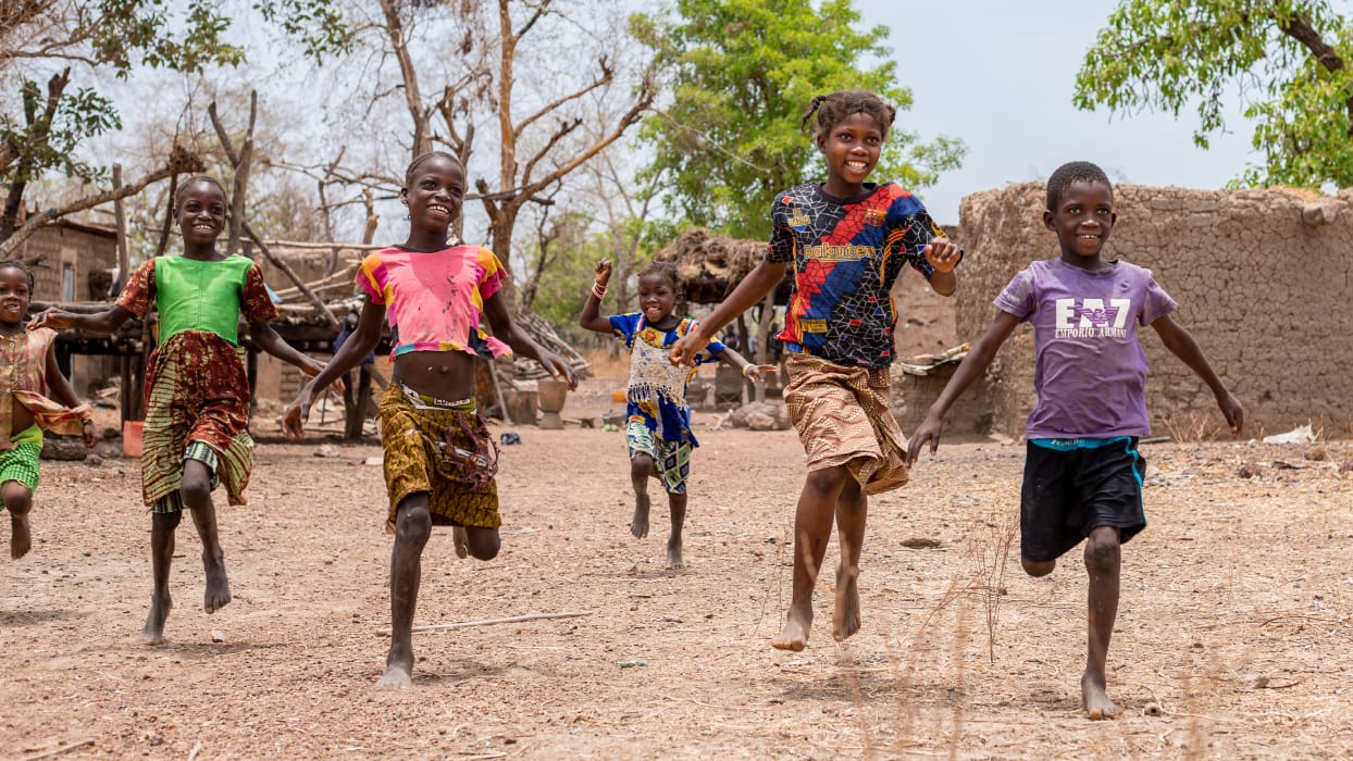 Smiling children running barefoot on a dirt path in a rural village under a clear sky.