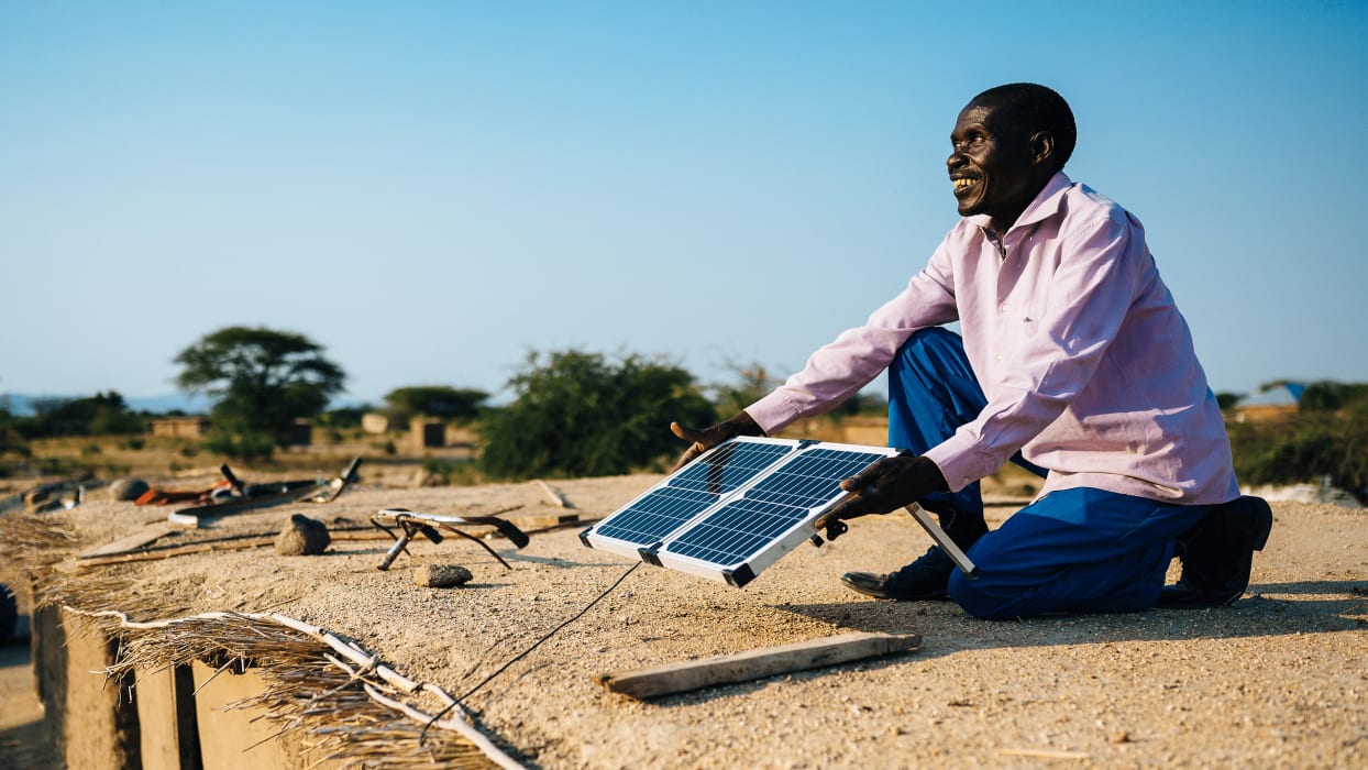 A man is knelt on top of a straw and mud roof. He holds a solar panel in his hands whilst looking up and smiling.
