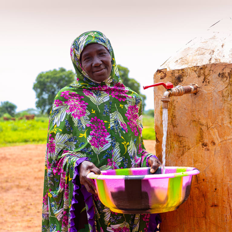 Smiling woman wearing a green and pink floral patterned outfit, holding a multicoloured basin under a flowing outdoor water tap. A small house and trees are visible in the rural background.