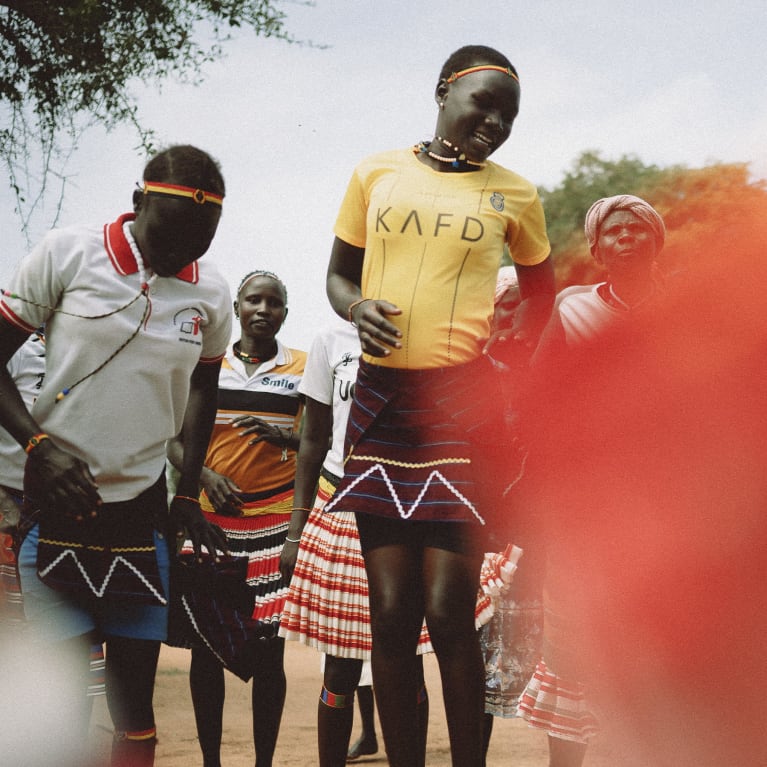 Group of people in traditional and modern attire with beaded skirts and headbands, prominent red blur.