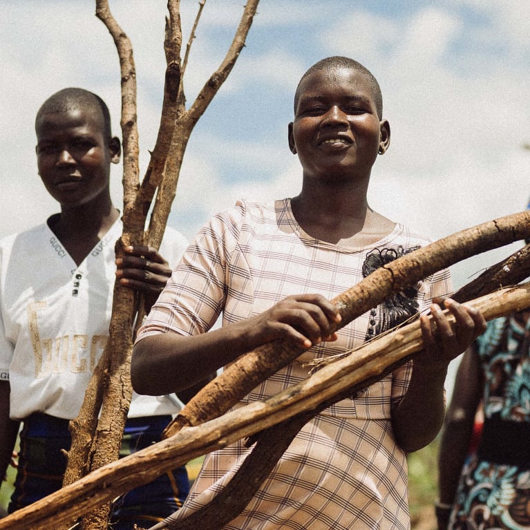 A young woman with a shaved head smiles, holding several thick branches, alongside a second young person holding a branched stick. Other blurred figures and green plants are in the background under a cloudy sky.