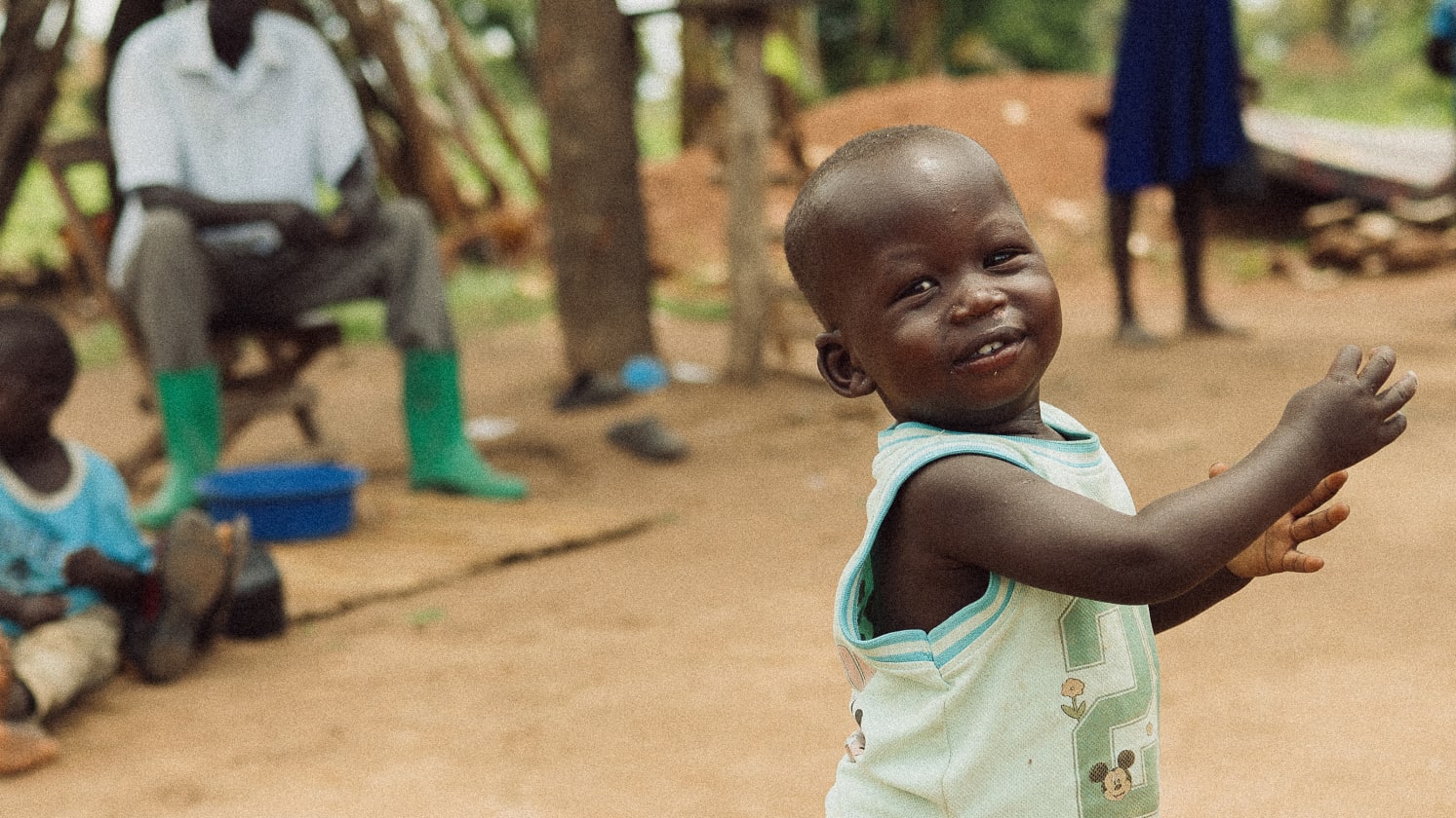 A smiling child in a light shirt looks towards the camera, with blurred people in the background.