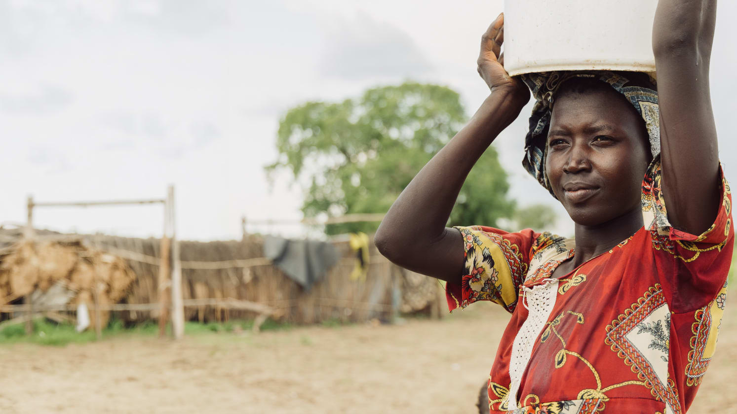 A woman in a red patterned dress and headwrap carries a white bucket on her head, looking ahead. The blurred background shows a dry, rural area with simple structures and a distant tree.