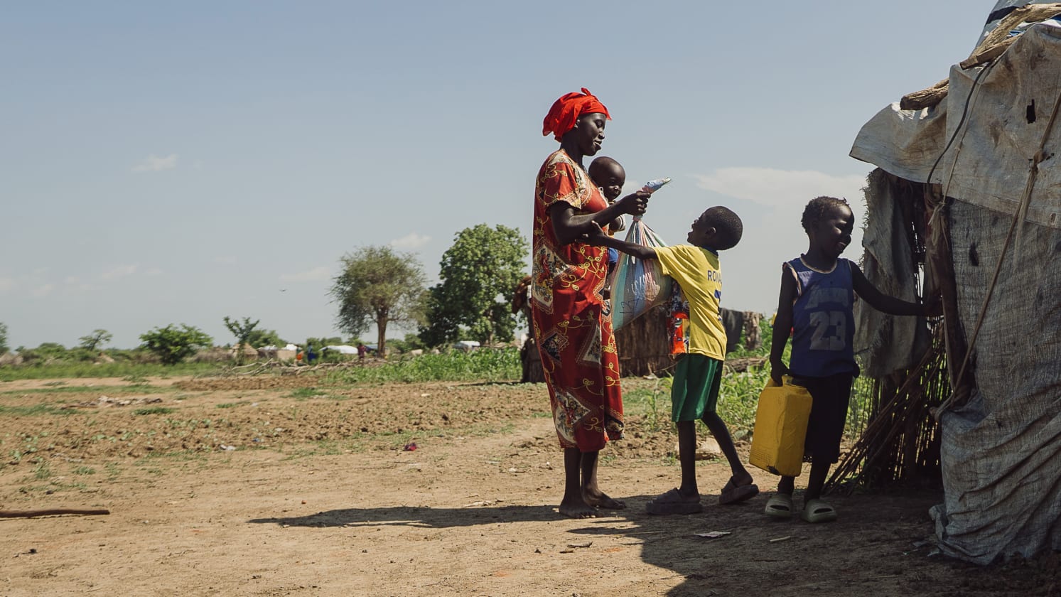 A woman in a red headscarf holds a baby and hands a bag to a boy in a yellow top. Another boy holding a yellow jerry can stands by a makeshift shelter. The scene is outdoors on dry, dusty ground under a clear sky.