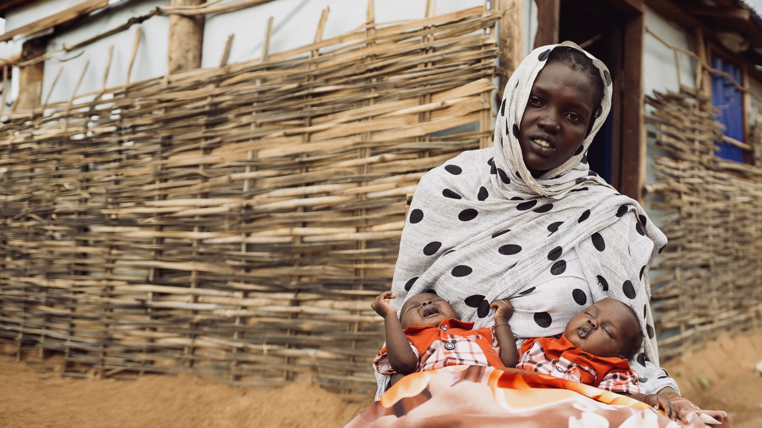 A woman in a white headscarf with black polka dots holds two infants, one crying and one sleeping, in front of a woven wooden fence and simple building.
