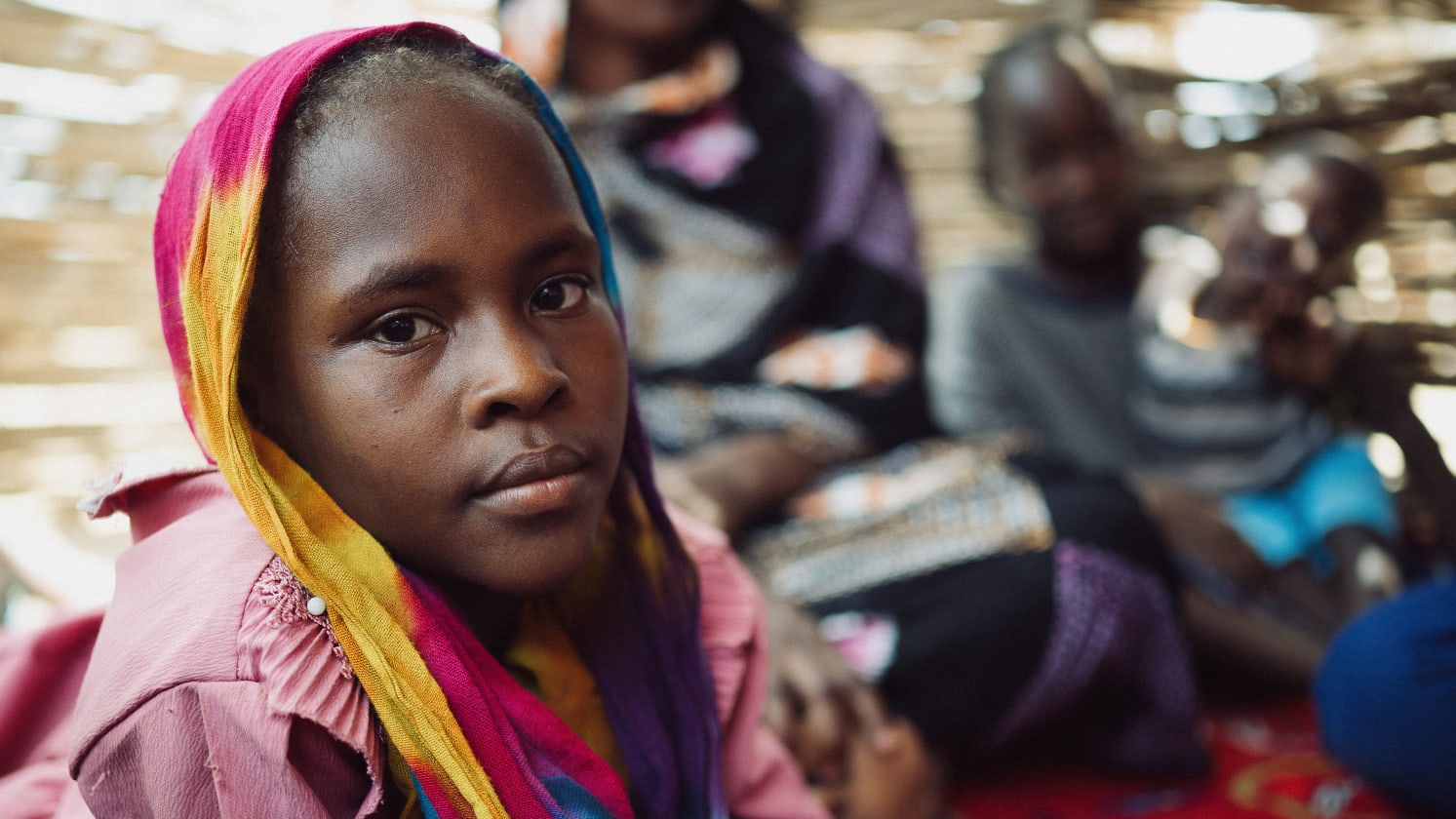 Close-up of a young Black girl with a colorful headscarf looking at the camera, blurred people behind.