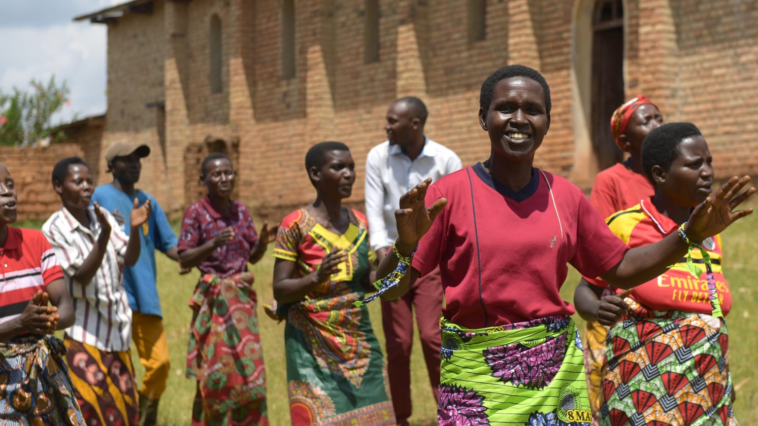 A group of people outside a brick building are singing and clapping with smiles.