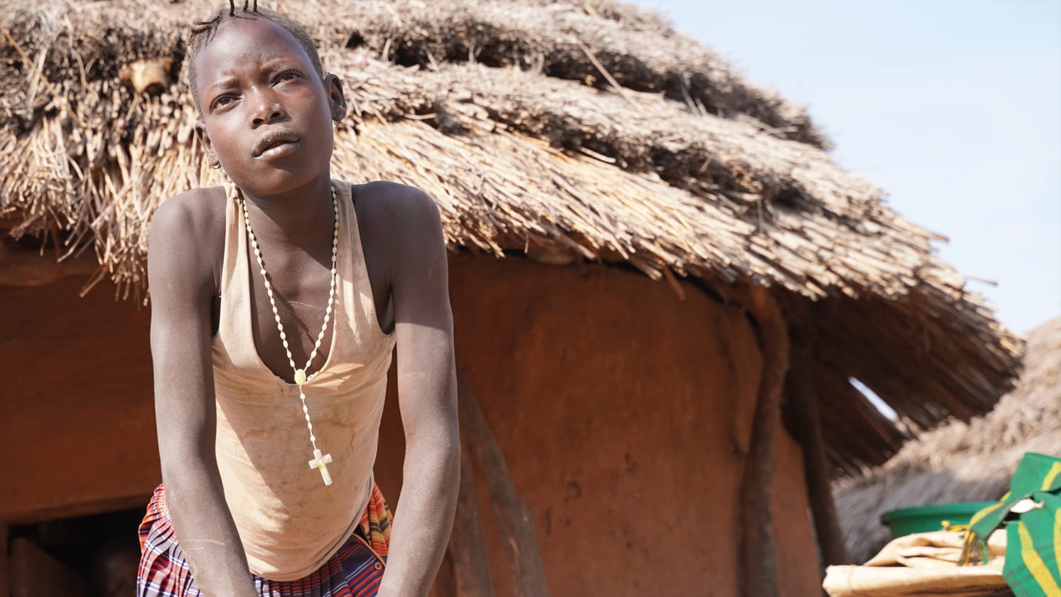 A person wearing a cross necklace leaning forward on their hands outside of a hut