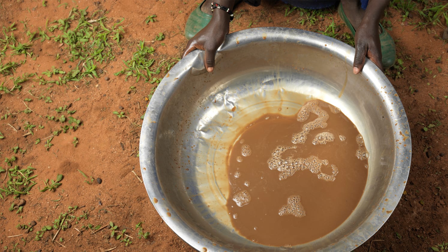 Person holds a metal basin with muddy water, standing on red earth.