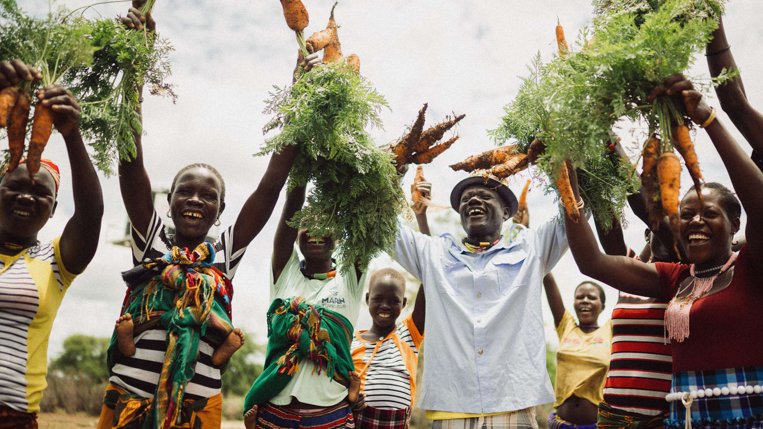 A joyful group of adults and children raise bunches of freshly harvested carrots with green tops above their heads.
